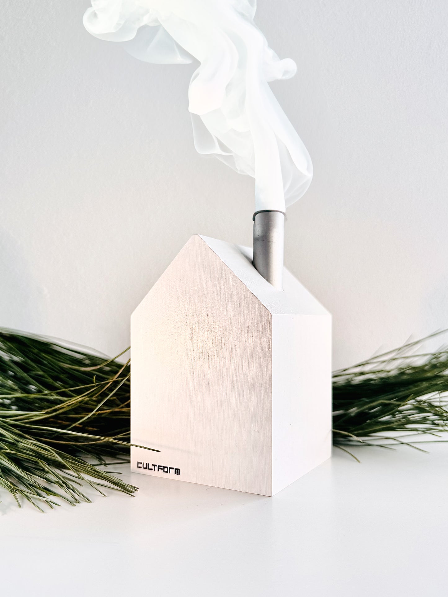 Weißes Räucherhaus aus Holz mit Rauch, das aus einem Schornstein auf der Dachspitze aufsteigt, umgeben von grünen Zweigen, Produktkomponente vom Genuss Set Chemnitz