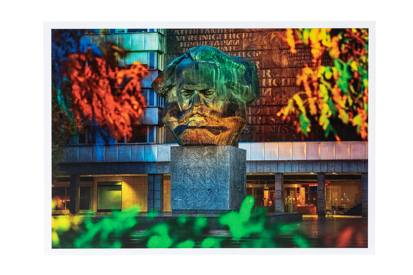 Postkarte mit Foto: Große steinerne Büste von Karl Marx vor einem Gebäude, umgeben von buntem Herbstlaub in Rot, Grün und Gelb