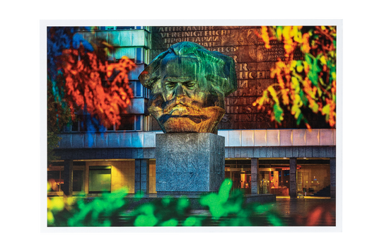 Postkarte mit Foto: Große steinerne Büste von Karl Marx vor einem Gebäude, umgeben von buntem Herbstlaub in Rot, Grün und Gelb